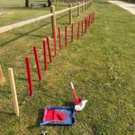 Image d'un chantier en plein air avec des piquets rouges récemment peints alignés le long d'une clôture en bois. On aperçoit également un seau de peinture rouge, un rouleau et un pinceau sur un plateau, suggérant un projet de peinture en cours. L'environnement est verdoyant, avec des arbres en arrière-plan et un chemin de terre adjacent, créant une ambiance paisible et naturelle. Cette image illustre des travaux de jardinage ou d'aménagement paysager
