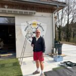 Un homme souriant se tient devant un bâtiment d'association sportive, tenant une échelle. Il porte un pull noir, un pantalon rouge et des chaussures de sport. À l'arrière-plan, on aperçoit le logo de l'association, ainsi que des équipements de jardinage et de bricolage. L'environnement est ensoleillé, avec des arbres en arrière-plan, ce qui suggère une journée agréable. Cette image illustre l'engagement communautaire et les activités de maintenance au sein d'une