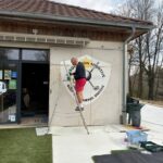 Un homme est en train de peindre un mur à l'extérieur d'un bâtiment associatif sportif, représentant un logo de golf. Il utilise une échelle pour atteindre la partie supérieure du mur, tandis que des outils de peinture et des fournitures sont visibles au sol. L'environnement est paisible, avec des arbres en arrière-plan et un espace de verdure aménagé. Cette activité souligne l'engagement de la communauté dans l'entretien et l'embellissement des installations sportives.
