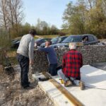Trois hommes travaillent ensemble sur un projet de construction en extérieur, avec un mur en gabions en arrière-plan. L'un d'eux est à genoux, utilisant des outils pour fixer des planches en bois sur une base en béton. Les autres observateurs se tiennent debout, discutant des étapes du projet. Des voitures sont garées à proximité, et l'environnement est verdoyant avec des arbres en arrière-plan sous un ciel bleu clair. Cette scène illustre le travail collaboratif