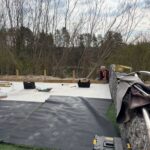 Travaux de construction en cours sur un espace extérieur, avec un ouvrier en train de préparer le sol. Le chantier présente une surface en béton partiellement couverte de tapis en caoutchouc noir et de gazon synthétique. En arrière-plan, on aperçoit une forêt avec des arbres dénudés, typiques d'une saison printanière. Des outils et équipements de construction sont visibles, témoignant de l'avancement des travaux. L'image illustre l'aménagement d'un