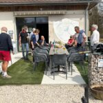 Groupe de personnes se rassemblant devant un bâtiment, probablement un club ou une association. La scène se déroule à l'extérieur, avec une grande table et des chaises en rotin noir. Des boissons et des collations sont visibles sur la table. L'environnement est convivial, avec une pelouse synthétique et un espace en gravier. L'arrière-plan montre un mur avec le logo d'une association sportive, suggérant une activité de loisir ou de détente.