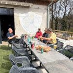 Groupe d'hommes se réunissant autour d'une table en terrasse, profitant d'un moment convivial au golf des Quatre Saisons. L'image montre des boissons variées sur la table, des baguettes et un logo de l'association sur le mur. L'environnement extérieur est apaisant, avec des arbres en arrière-plan, suggérant une atmosphère détendue et amicale propice aux échanges après une partie de golf.