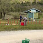 Deux personnes, une femme et un homme, s'affairent à préparer un terrain de jeu en plein air, avec des piquets rouges plantés dans le sol. La femme est debout, tandis que l'homme est accroupi près d'un seau rouge. En arrière-plan, un abri en bois avec une enseigne "Bienvenue" est visible, entouré d'une verdure printanière. Des paniers en plastique verts sont empilés à côté d'un seau