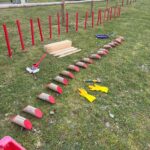 Image illustrant un projet de bricolage en extérieur, mettant en avant des poteaux en bois peints en rouge disposés sur une pelouse. On peut voir des planches de bois, des outils de peinture, et des gants jaunes, suggérant une activité de construction ou de décoration. L'environnement est verdoyant, avec un chemin et des arbres en arrière-plan, indiquant un cadre naturel propice aux travaux manuels. Cette image peut inspirer des idées pour des projets de jardin