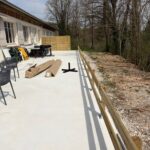 Vue d'une terrasse récemment aménagée avec du mobilier de jardin, incluant des chaises et des tables. Des matériaux de construction sont visibles, indiquant des travaux en cours. À l'arrière-plan, une clôture en bois délimite l'espace et des arbres dépouillés de leurs feuilles se dressent le long de la bordure. Cette image illustre un espace extérieur propice à la détente et aux activités en plein air, idéal pour profiter de la nature environnante.