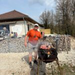 Homme en train de griller des viandes sur un barbecue en plein air, portant un t-shirt orange et une casquette blanche. En arrière-plan, une maison avec un mur en pierre et des arbres dénudés. Le barbecue dégage de la fumée, tandis que des convives se rassemblent pour profiter d'un moment convivial. Cette image évoque des activités de plein air, des barbecues et des rassemblements familiaux ou amicaux.