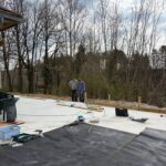 Deux personnes travaillent sur un projet de construction en extérieur, sur une surface en béton. Des outils tels qu'une scie et une perceuse sont visibles, ainsi que des matériaux comme des planches de bois et des rouleaux de revêtement noir. En arrière-plan, des arbres dénudés indiquent une période printanière. L'image illustre les étapes de la rénovation ou de l'aménagement d'un espace extérieur, idéal pour des projets de bricolage ou d'amélioration de l'hab