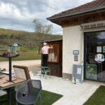 Une personne en tenue décontractée, perchée sur une chaise, peint la porte en bois d'un bâtiment situé à proximité d'un terrain de golf. L'environnement est verdoyant, avec des arbres en arrière-plan et un ciel nuageux. Des chaises et tables sont visibles sur le sol en béton, ajoutant une ambiance conviviale à l'espace extérieur. L'image illustre une activité de rénovation dans un cadre naturel, idéal pour les amateurs de golf et de loisirs en plein air.