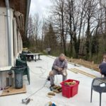 Un homme travaille sur un projet de bricolage à l'extérieur d'un bâtiment, assis sur une chaise à proximité d'une boîte rouge contenant des pièces en bois. Il utilise un outil électrique pour façonner des éléments en bois, tandis que d'autres personnes s'affairent en arrière-plan, à proximité d'un espace de travail. L'environnement est naturel, avec des arbres dénudés en arrière-plan, suggérant une saison printanière. Des matériaux, tels que des planches de bois