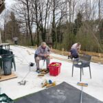 Travaux de rénovation extérieure en cours avec plusieurs personnes s'affairant sur un chantier. L'image montre un espace de patio en béton où des membres d'une équipe de construction s'occupent de la mise en place de structures en bois. On peut voir des outils de bricolage, un seau rouge rempli de matériaux, et des chaises en plastique. En arrière-plan, des arbres dénudés ajoutent une touche naturelle à l'environnement. Cette scène illustre l'importance du travail