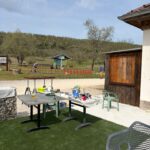 Un espace extérieur accueillant avec des tables de travail et des fournitures de peinture, situé près d'un chemin gravelé. En arrière-plan, des collines verdoyantes et une signalisation de loisirs. L'environnement est propice aux activités de plein air, idéal pour les projets artisanaux ou les réunions conviviales. Le mobilier inclut des chaises confortables et un verre sur la table, suggérant un moment de détente.