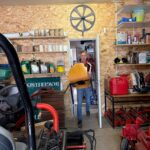 Un homme portant un objet en bois entre dans un garage bien organisé, où divers outils de jardinage et produits d'entretien sont visibles sur les étagères. Le garage est décoré de panneaux et d'une roue en bois au mur. On aperçoit également une tondeuse et d'autres équipements de jardinage au premier plan. L'espace est lumineux, avec des murs en contreplaqué et un sol en béton. Cette image illustre un environnement de travail pratique pour les passionnés de jardin