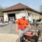 Un homme en t-shirt orange prépare un barbecue en extérieur, grillant des viandes variées telles que des saucisses et des côtes. En arrière-plan, un groupe de personnes discute près d'un bâtiment en pierre avec des fenêtres à volets. La scène se déroule par une journée ensoleillée, créant une ambiance conviviale et festive. Le barbecue est entouré de gravier, ajoutant un aspect rustique à l'événement.