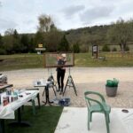 Une personne travaille sur un projet de peinture en plein air, tenant un cadre en bois sur des tréteaux. La scène est entourée de verdure et de collines, avec des signes directionnels indiquant un parcours à proximité. Une table est chargée de fournitures de peinture, tandis qu'une chaise en plastique vert est visible au premier plan. L'atmosphère est calme et propice à la créativité. Cette image illustre une activité artistique en extérieur, mettant en avant le travail