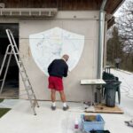 Un homme peint un logo sur un mur extérieur d'un bâtiment sportif, représentant l'Association Sportive. Il utilise une échelle pour atteindre la partie supérieure du design. Des outils de peinture et des fournitures sont visibles au sol, tandis qu'un autre homme se trouve en arrière-plan. L'environnement est en plein air, avec des arbres en arrière-plan et un espace bien entretenu. Cette activité met en avant l'engagement communautaire dans le sport et l'amélioration des infrastructures.