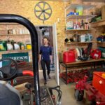 Une personne entre dans un garage bien organisé, où divers outils et équipements de jardinage sont visibles. L'espace est doté de rangements en bois, contenant des produits d'entretien et des accessoires pour l'entretien des espaces extérieurs. En arrière-plan, un panneau mural et des équipements de jardinage sont soigneusement disposés, soulignant l'aspect fonctionnel du lieu. L'éclairage est adéquat, permettant une bonne visibilité dans cet environnement de travail.
