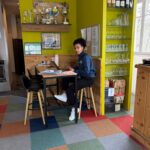 Une personne assise à une table dans un espace de travail moderne, entourée de décorations vibrantes. Le mur est peint en jaune vif et orné de trophées et de médailles. Des bouteilles de vin sont disposées sur une étagère à droite, tandis que le sol est recouvert de dalles colorées, ajoutant une touche dynamique à l'environnement. L'espace est lumineux et accueillant, parfait pour le travail ou les études.