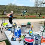 Une femme est en train de peindre un meuble en bois dans un environnement extérieur, entourée de matériel de bricolage. Sur une table, on peut voir plusieurs pots de peinture, des pinceaux et d'autres outils de décoration. En arrière-plan, le paysage verdoyant est parsemé d'arbres et d'une structure en bois, tandis que d'autres personnes s'affairent à des activités similaires. Cette scène évoque le travail manuel et la créativité dans un cadre naturel.