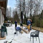 Groupe de personnes travaillant sur une terrasse extérieure, entouré d'un environnement naturel. Des tables et des chaises sont disposées sur le sol, tandis que quelques individus discutent autour d'un parasol. L'activité semble liée à l'aménagement ou à la rénovation de l'espace extérieur. Des outils sont visibles, indiquant un projet de bricolage en cours. Les arbres environnants sont dépouillés, suggérant une période de transition saisonnière.