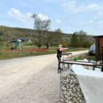 Activité en plein air dans un cadre naturel, avec des participants engagés dans des tâches variées. On aperçoit une femme en train de manipuler des objets sur une table, tandis qu'une autre femme prépare du matériel à proximité. En arrière-plan, un abri en bois et une route en gravier serpentent à travers un paysage verdoyant, avec des arbres et de la végétation printanière. Cette image évoque une ambiance conviviale et dynamique, idéale pour des événements communaut