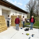Quatre hommes s'affairent à la construction d'une clôture en bois devant une maison. Deux d'entre eux tiennent des panneaux en bois, tandis qu'un autre ajuste les éléments de la structure. Un homme observe le travail en tenant des outils. L'environnement est calme, avec des arbres en arrière-plan et des matériaux de construction visibles. L'image illustre un projet de bricolage collaboratif, mettant en avant l'esprit d'équipe et l'importance de l'entretien extérieur des habitations