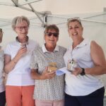 Quatre femmes souriantes se tiennent ensemble sous un chapiteau, chacune tenant un petit pot ou un cadeau. Elles portent des polos blancs, ce qui suggère une activité de groupe ou un événement. En arrière-plan, des décorations colorées et des tables sont visibles, ajoutant une ambiance festive. Cette image pourrait illustrer une cérémonie de remise de prix ou une rencontre conviviale.