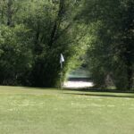 Vue d'un parcours de golf verdoyant avec un drapeau blanc signalant le trou. En arrière-plan, des arbres feuillus entourent une étendue d'eau, créant un cadre naturel apaisant. La lumière du soleil éclaire le terrain, accentuant la beauté du paysage. Cette image évoque la tranquillité et le plaisir du golf en pleine nature.