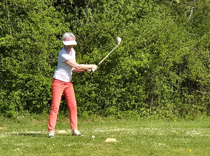 Femme en train de jouer au golf sur un parcours verdoyant, vêtue d'un polo blanc et d'un pantalon rouge. Elle se prépare à frapper la balle avec un club de golf, sous un ciel ensoleillé. En arrière-plan, une végétation dense ajoute une touche naturelle à la scène. Cette image illustre l'activité sportive et le plaisir du golf en plein air.