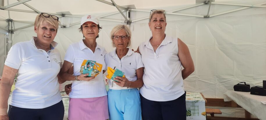 Quatre femmes souriantes posent ensemble sous un chapiteau lors d'un événement. Elles portent des polos blancs et tiennent des petits paquets colorés, probablement des récompenses ou des souvenirs. L'ambiance est festive et conviviale, reflétant un moment de célébration ou de compétition amicale. En arrière-plan, on aperçoit une table avec divers articles, suggérant un événement communautaire ou sportif. Cette image illustre l'esprit d'équipe et l'engagement dans