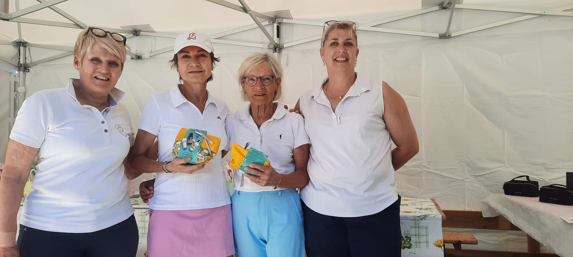 Quatre femmes souriantes posent ensemble sous un chapiteau lors d'un événement. Elles portent des polos blancs et tiennent des petits paquets colorés, probablement des récompenses ou des souvenirs. L'ambiance est festive et conviviale, reflétant un moment de célébration ou de compétition amicale. En arrière-plan, on aperçoit une table avec divers articles, suggérant un événement communautaire ou sportif. Cette image illustre l'esprit d'équipe et l'engagement dans