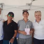 Quatre femmes souriantes posent ensemble sous un chapiteau, vêtues de polos de golf. Elles semblent participer à un événement convivial, probablement lié au golf, avec des récompenses en arrière-plan. L'une d'elles tient un sac rose, suggérant qu'il pourrait s'agir d'un prix ou d'un cadeau. Cette image illustre l'esprit de camaraderie et de compétition amicale souvent présent dans les tournois de golf.