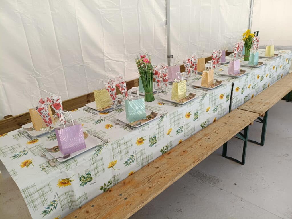 Table décorée pour un événement festif, avec une nappe à motifs floraux et des assiettes disposées soigneusement. Des sacs cadeaux colorés sont placés sur chaque assiette, ajoutant une touche festive à la décoration. Des bouquets de fleurs complètent l'ambiance accueillante. Cette mise en scène est idéale pour des célébrations telles que des anniversaires, des mariages ou des repas de famille.