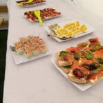 Table de buffet avec diverses préparations culinaires. À gauche, des verrines de crevettes et de salade, à côté de bouchées de saumon fumé garnies de légumes frais. Au centre, des œufs mimosa décorés de crème jaune. À l'arrière-plan, des plateaux de fruits et charcuterie colorés, accompagnés de pinces vertes pour le service. Idéal pour un événement festif ou un apéritif.